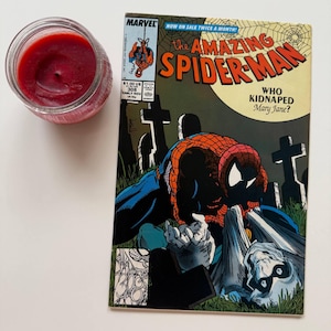 Könnte beinhalten: Eine Kerze im Glas mit tiefrotem Wachs liegt neben einem Comic. Das Comic-Cover zeigt Spider-Man in einer Friedhofsszene mit dem Titel "The Amazing Spider-Man" und dem Text "Who Kidnapped Mary Jane?"
