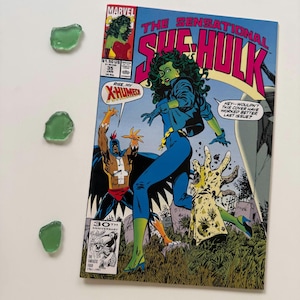 May include: A comic book titled "The Sensational She-Hulk" featuring a green-skinned female superhero in a blue outfit. The cover art includes the Marvel logo and the text "Rise, My X-Humed". Three pieces of sea glass are also present.