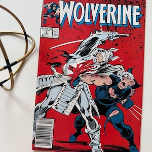 Könnte beinhalten: Ein Cover des Marvel Comics Wolverine #2, auf dem Wolverine in einem rot-weißen Samurai-Kostüm gegen einen anderen Wolverine in einem blau-schwarzen Kostüm kämpft. Das Cover ist rot mit dem Titel "Wolverine" in weißen und blauen Buchstaben.