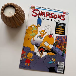Könnte beinhalten: Ein brauner, strukturierter Kerzenhalter mit einer weißen Kerze neben einem Simpsons Comics #1 Comic. Das Comic-Cover zeigt Homer Simpson mit dem Text "THE AMAZING COLOSSAL HOMER!"