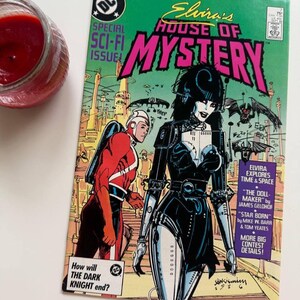 Peut inclure: Une couverture de bande dessinée mettant en scène Elvira, la maîtresse des ténèbres, vêtue d'une tenue noire et argentée, debout devant une ville futuriste. Le titre de la bande dessinée est "Elvira's House of Mystery" et la couverture présente le texte "Special Sci-Fi Issue".