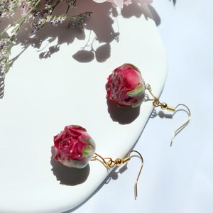 May include: A pair of gold earrings with a pink rose bud encased in resin. The earrings are on a white surface.