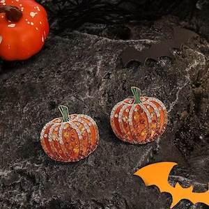 May include: Two orange pumpkin-shaped brooches with green stems and encrusted with rhinestones. The brooches are on a dark, textured surface with a small orange pumpkin and a bat-shaped cutout.