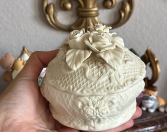 May include: Cream-colored, decorative trinket box with a floral design. The lid features a cluster of roses and leaves. The box has a textured, basketweave pattern and floral relief details. The box is held in a hand.