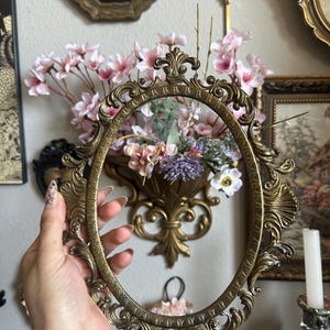 May include: An antique brass oval picture frame with ornate detailing is held in front of a wall with other vintage decor. The frame is empty, showcasing its intricate design. Pink and purple flowers are visible in the background.