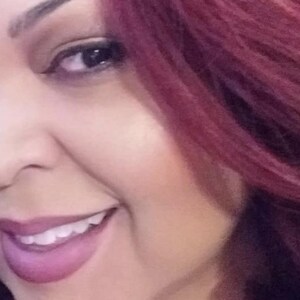 May include: A close-up of a woman's face with long, dark red hair. She is smiling and her teeth are visible. Her makeup is natural and she is wearing a light pink lipstick.