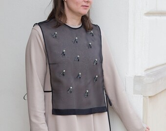 Op de afbeelding: Een vrouw draagt een lange, beige jurk met een zwart mesh vest met een patroon van kleine, zwarte strikjes. Het vest heeft een zwarte rand rond de halslijn en onderkant.
