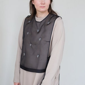 Pode incluir: Uma mulher vestindo um vestido bege de manga comprida com um colete de tule preto sobreposto com detalhes decorativos.