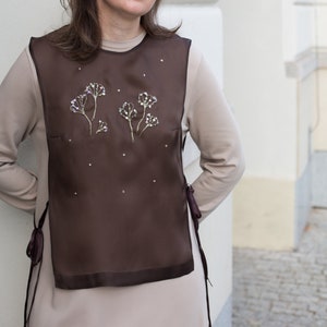 Pode incluir: Um top marrom sem mangas com uma sobreposição transparente que apresenta desenhos florais bordados. A sobreposição tem um laço nas laterais.