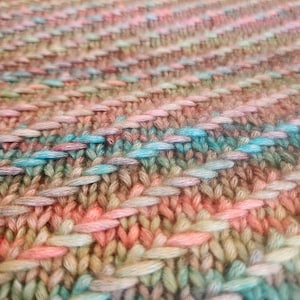 May include: Close-up of a hand-knitted textile with a textured surface. The yarn is a blend of colors, including peach, light blue, and green. The stitches create a pattern of horizontal and diagonal lines, giving the fabric a unique, handcrafted appearance.