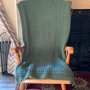 May include: A green knitted throw blanket draped over a wooden rocking chair. The blanket has a waffle stitch pattern.