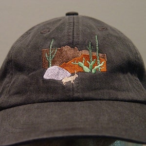 Könnte beinhalten: Eine dunkelgraue Baseballkappe mit einem gestickten Design einer Wüstenlandschaft mit Kakteen und einem kleinen Tier.