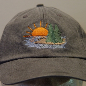 May include: A dark grey baseball cap with an embroidered design of a sunset over a small island with two pine trees.