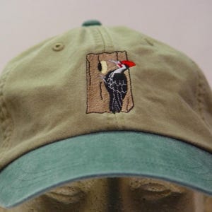 Puede incluir: Gorra de béisbol caqui con visera y botón superior verdes. La gorra presenta una imagen bordada de un pájaro carpintero blanco y negro con cabeza roja sobre fondo de madera marrón.