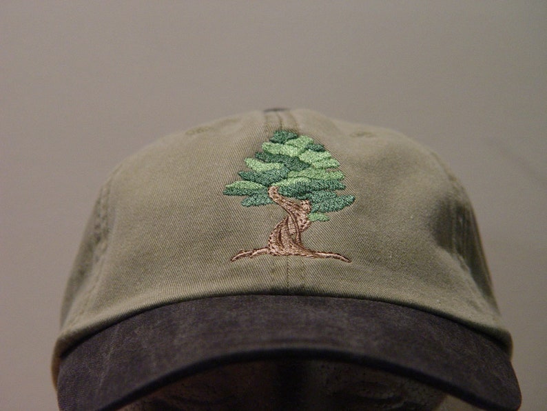 May include: A green and brown embroidered tree design on a khaki baseball cap with a black brim.