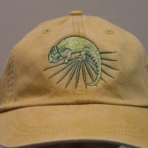 Può includere: Un cappello da baseball color beige con un'immagine ricamata di un camaleonte verde su una pianta verde.
