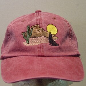 May include: A red baseball cap with an embroidered design of a coyote howling at the moon in a desert landscape with a cactus and a rock formation.