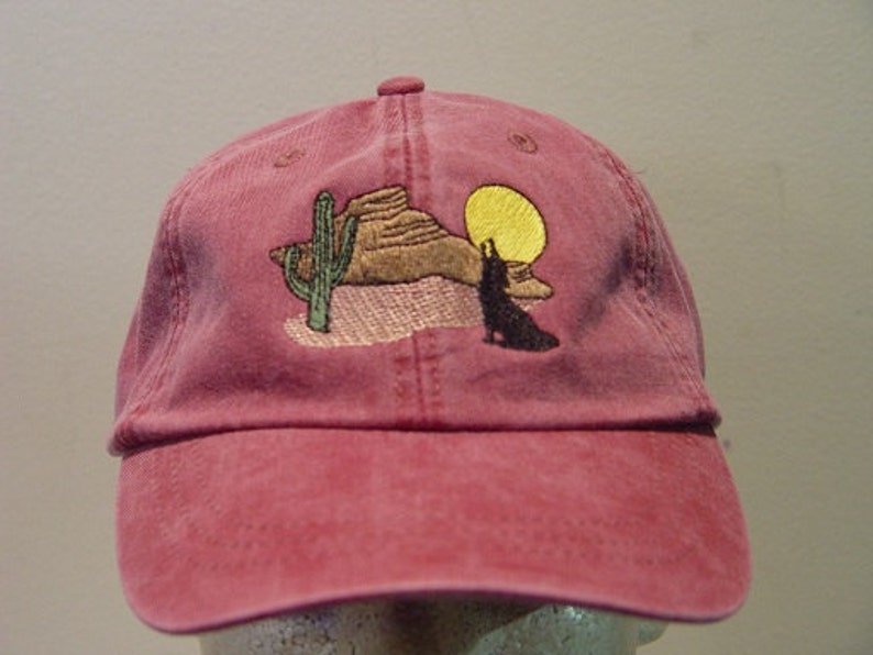 May include: A red baseball cap with an embroidered design of a coyote howling at the moon in a desert landscape with a cactus and a rock formation.