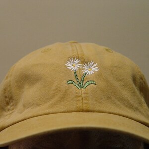 May include: A tan baseball cap with an embroidered design of two white daisies with green stems.
