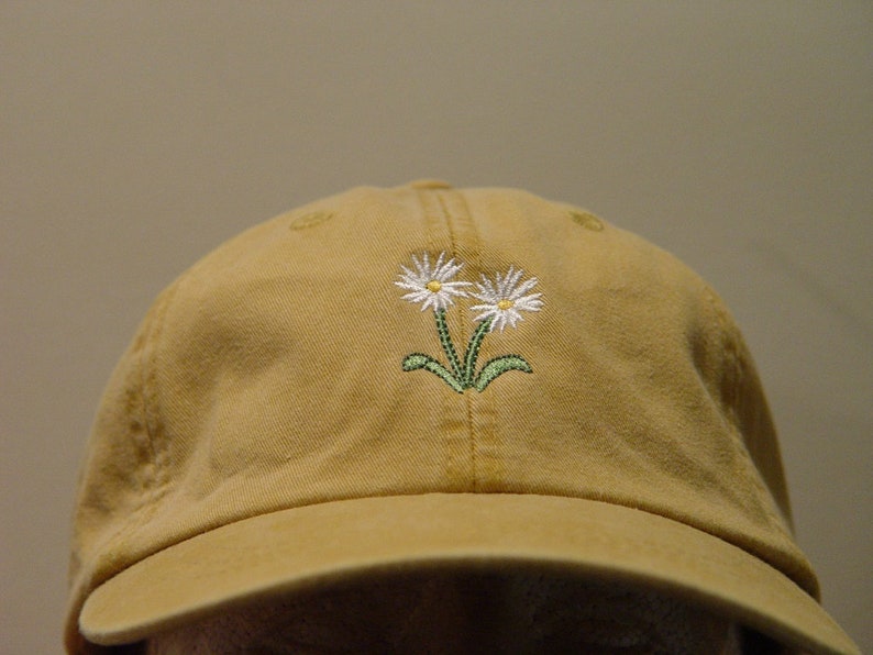 May include: A tan baseball cap with an embroidered design of two white daisies with green stems.