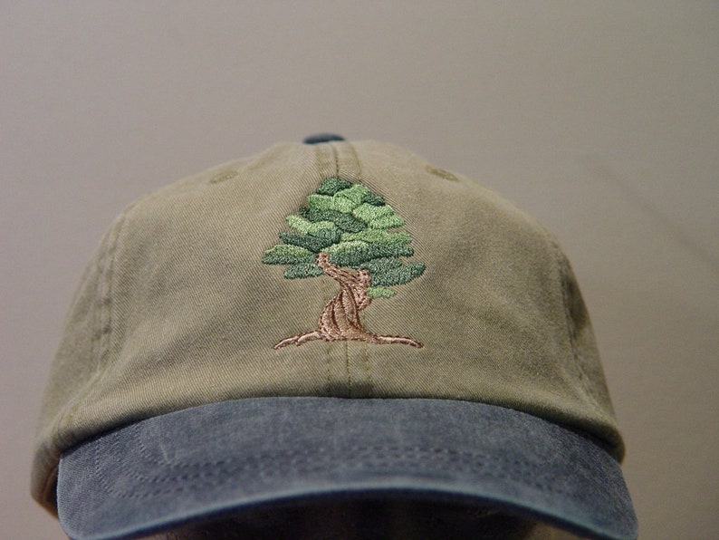 May include: A green and brown embroidered tree design on a khaki baseball cap with a blue brim.