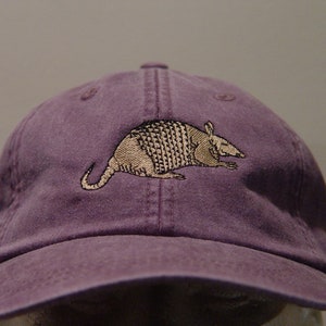 May include: A purple baseball cap with an embroidered armadillo design in brown and beige.