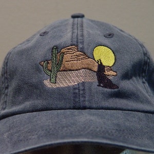 May include: A blue denim baseball cap with an embroidered design of a desert scene with a cactus, a mountain, a coyote, and a yellow sun.