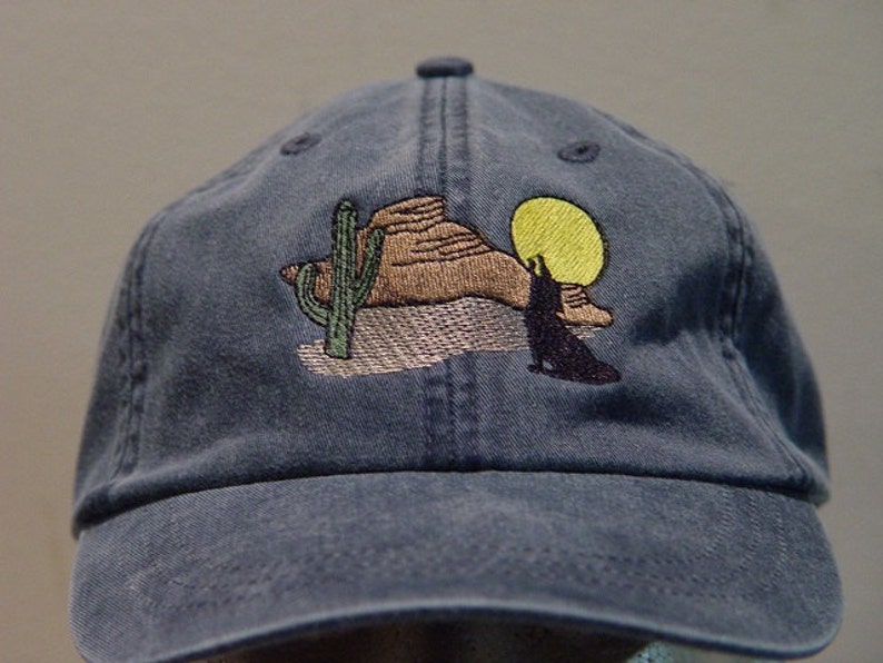 May include: A blue denim baseball cap with an embroidered design of a desert scene with a cactus, a mountain, a coyote, and a yellow sun.