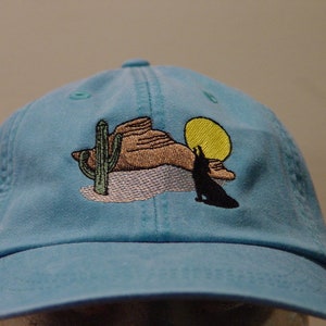 May include: A light blue baseball cap with an embroidered design of a desert scene with a cactus, a mountain, a coyote, and a yellow sun.