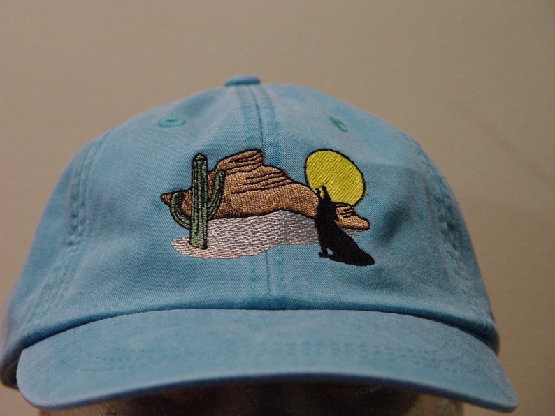 May include: A light blue baseball cap with an embroidered design of a desert scene with a cactus, a mountain, a coyote, and a yellow sun.