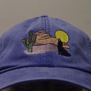 May include: A blue baseball cap with an embroidered design of a desert scene with a cactus, a mountain, a wolf howling at the moon, and a yellow sun.