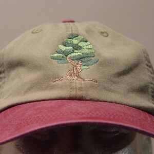 May include: A khaki baseball cap with a red brim and an embroidered green tree design.