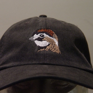 Op de afbeelding: Een zwarte baseballpet met een geborduurd beeld van een bruine en witte vogel met een rode kroon.