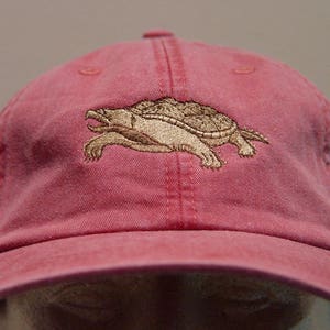 Pode incluir: Boné de beisebol rosa desbotado com aba curva. O boné apresenta um design bordado detalhado de uma tartaruga em tons de bege e castanho. O boné é feito de um tecido macio e desgastado.