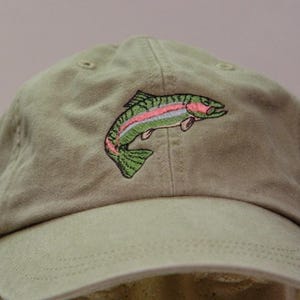 以下が含まれることがあります： 跳ねるニジマスが刺繍されたカーキ色のベースボールキャップ。魚は緑、ピンク、白の糸で表現されており、キャップのフロントパネルにはこの種の特徴である側線がはっきりと描かれています。