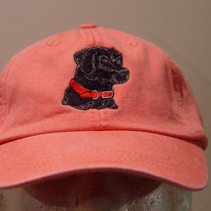 Pu&ograve; includere: Un berretto da baseball in cotone rosa con un Labrador Retriever nero ricamato che indossa un collare rosso.