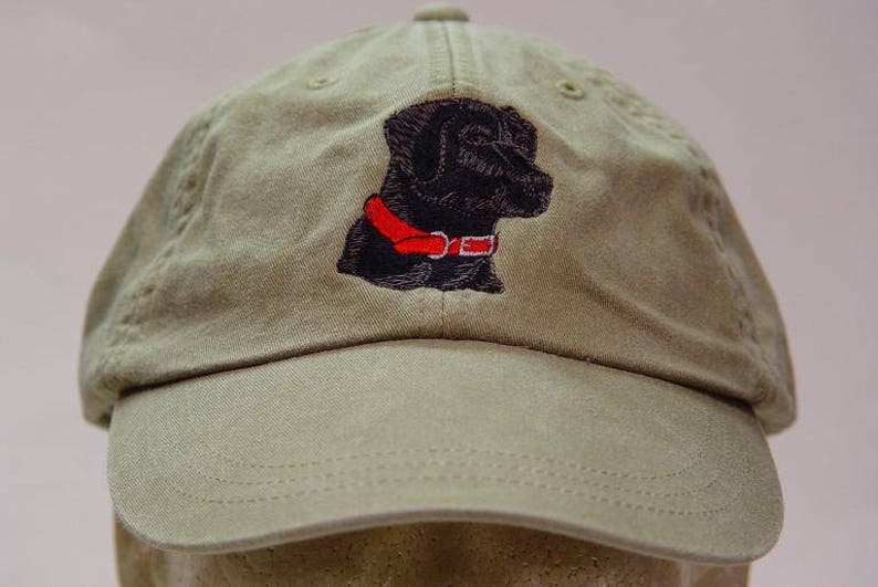 Pu&ograve; includere: Un cappello da baseball verde kaki con un labrador retriever nero ricamato che indossa un collare rosso.