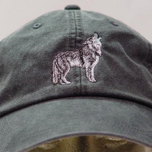 May include: A charcoal gray baseball cap featuring an embroidered design of a wolf standing in profile. The cap has a curved brim, a structured front panel, and stitched eyelets on the crown, showcasing a detailed white and gray thread wolf graphic.