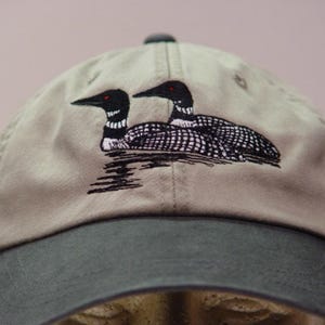 May include: A tan baseball cap with a dark green bill and top button. The cap features an embroidered design of two loons on water, with black and white detailing and red eyes. The loons are a prominent feature on the front of the cap.