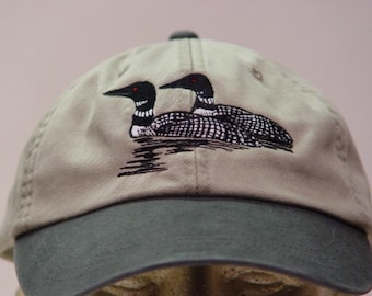 May include: A tan baseball cap with a dark green bill and top button. The cap features an embroidered design of two loons on water, with black and white detailing and red eyes. The loons are a prominent feature on the front of the cap.