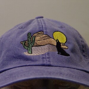 May include: A purple baseball cap with an embroidered design of a desert scene with a cactus, a mountain, a sun, and a wolf howling at the moon.