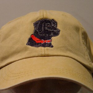 Pu&ograve; includere: Un cappello da baseball in tela beige con un Labrador Retriever nero ricamato che indossa un collare rosso.