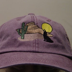 May include: Purple baseball cap with an embroidered design of a wolf howling at the moon, a cactus, and a mountain range.