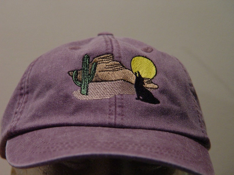 May include: Purple baseball cap with an embroidered design of a wolf howling at the moon, a cactus, and a mountain range.
