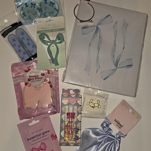 May include: A collection of beauty and hair accessories including blue eye pads, a green claw clip, a pink bow-shaped bath bomb, a bottle of holographic glow drops, gold-toned bow earrings, a light blue satin scrunchie, and a white notebook with blue bows.