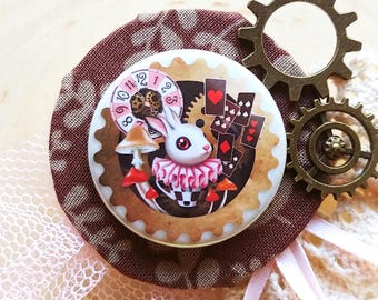 May include: A whimsical brooch featuring a white rabbit with a clock and playing cards, set against a brown background. The design includes bronze gears, a pink net, and lace trim, creating a steampunk aesthetic.