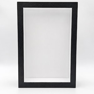 Puede incluir: Un marco tipo caja de sombra rectangular con un borde exterior negro texturizado y una profundidad interior blanca lisa. El marco está orientado verticalmente, proporcionando un espacio profundo y abierto con bordes limpios y definidos.