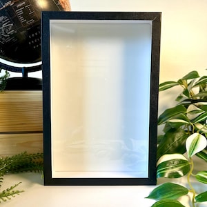 May include: A black picture frame with a white mount and a clear glass front. The frame is sitting on a wooden surface.