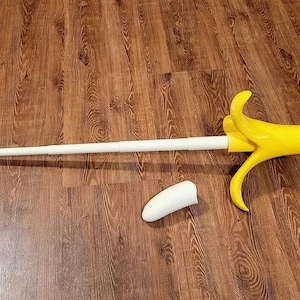 Könnte beinhalten: Ein gelbes bananenförmiges Schwert mit einem weißen Griff und einer weißen Bananenschale auf einem Holzboden.
