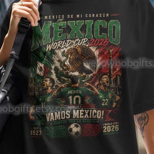 May include: A black t-shirt featuring a graphic print with the text MEXICO DE MI CORAZON, MEXICO WORLD CUP 2026, VAMOS MEXICO, and 1927 2026. The design includes illustrations of football players, a football, and the Mexican flag colours.